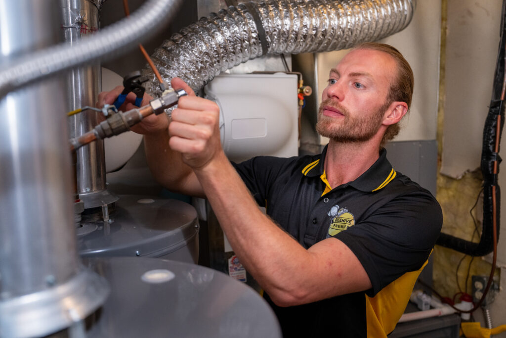 Image of Technician Working on a Water Heater – A Beehive Plumbing technician in a black and yellow uniform is adjusting a component on a water heater in a utility room. The background features pipes, ductwork, and insulation.