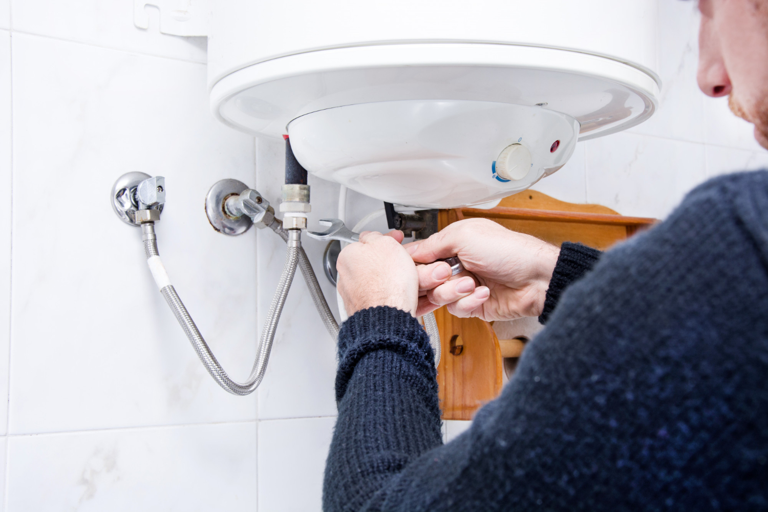 A man works on connecting water supply lines to a wall-mounted water heater using a wrench. The image emphasizes routine plumbing maintenance in a tiled bathroom.