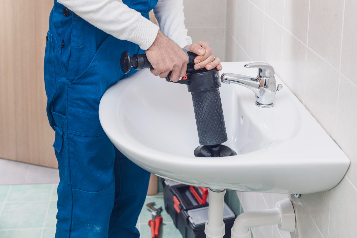 A plumber in blue overalls uses a powerful plunger tool to unclog a white bathroom sink. The surrounding tools suggest an active repair job in progress.