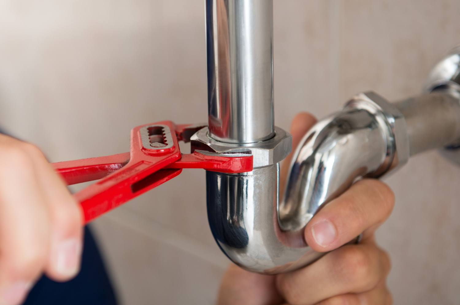 A person uses a red pipe wrench to tighten a shiny metal pipe connection, focusing on plumbing work. The close-up shot highlights the precision and tools used for pipe maintenance.